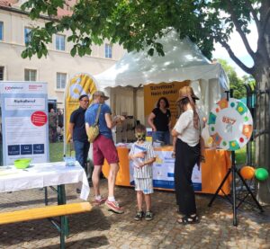 Es ist ein bunter Infostand des Projekts zu sehen, davor stehen mehrere Personen.