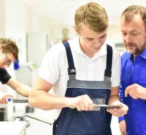 Blick in eine Werkstatt. Vorn steht ein junger Mann in Arbeitshose und hat ein Werkstück in den Händen. Ein älterer Mann im Blaumann erklärt ihm was an dem Werkstück. Im Hintergrund steht ein weiterer junger Mann und feilt etwas.