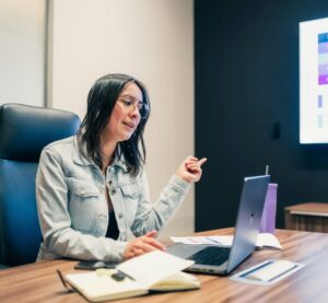 Eine Frau sitzt am Laptop und erzählt in einem Meeting etwas. Neben dem Laptop liegt ein Notizbuch.