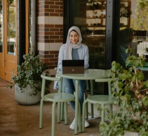 Eine Frau mit Kopftuch sitzt an einem kleinen Tisch in einem Kaffee an einem Laptop.