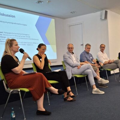Blick auf die Bühne. Mehrere Personen sitzen und zwei von ihnen sprechen ins Mikrofon.