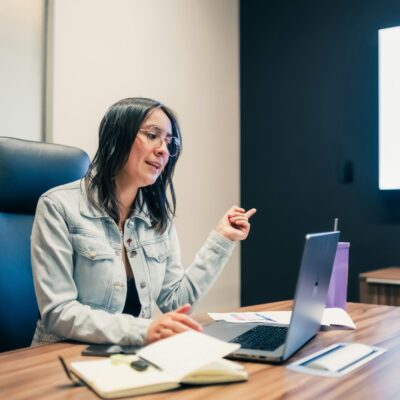 Eine Frau sitzt am Laptop und erzählt in einem Meeting etwas. Neben dem Laptop liegt ein Notizbuch.