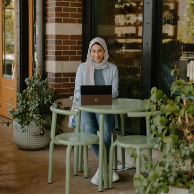 Eine Frau mit Kopftuch sitzt an einem kleinen Tisch in einem Kaffee an einem Laptop.