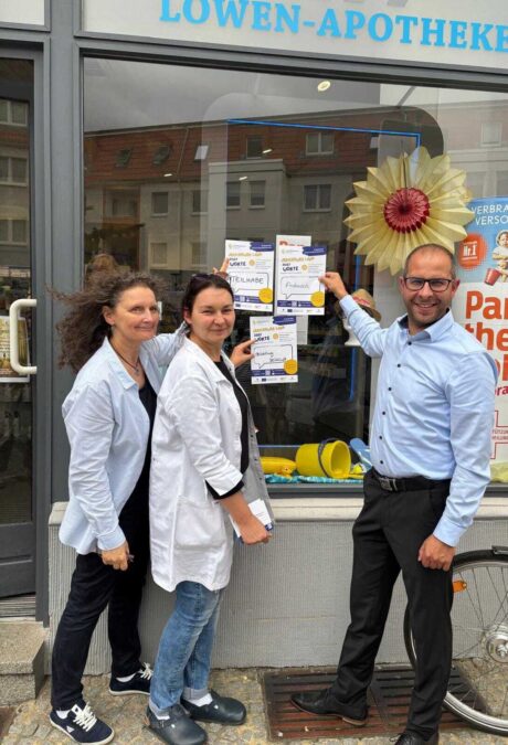 Vor einer Apotheke stehen zwei Apothekerinnen und der Bürgermeister der Stadt Burg, sie zeigen alle drei auf ein Poster, dass im Schaufenster hängt.