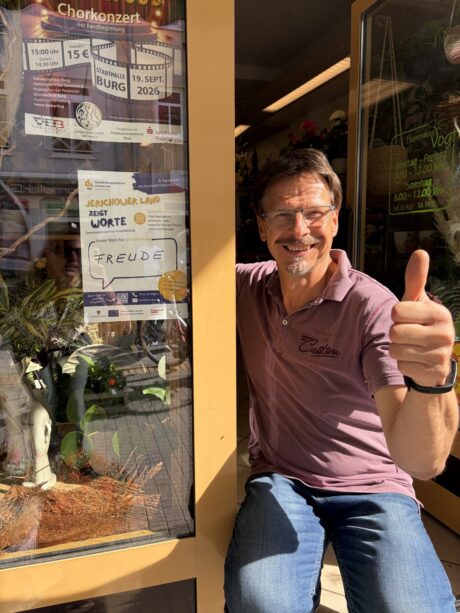 Ein Mann hockt im Eingangsbereich eines Geschäfts. Neben ihm ist das Schaufenster und dort hängt ein Plakat vom GBZ. Der Mann lächelt und hat den Daumen nach oben gehoben.