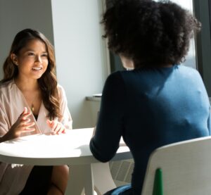 Zwei Frauen sitzen an einem Tisch und unterhalten sich. Es sieht nach einem Bewerbunsggespräch aus.