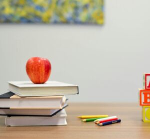Links ist ein Bücherstapel gekrönt mit einem Apfel. In der Mitte liegen ein paar Stifte und rechts sind drei Bauklötze mit den Buchstaben A, B und C gestapelt.