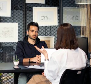 Eine Frau sitzt an einem Tisch und hat einen Laptop vor sich. Ihr gegenüber sitzt ein Mann. Es scheint, dass die Frau ihm Fragen stellt und er Bewerber für einen Job ist.