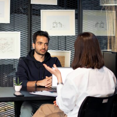 Eine Frau sitzt an einem Tisch und hat einen Laptop vor sich. Ihr gegenüber sitzt ein Mann. Es scheint, dass die Frau ihm Fragen stellt und er Bewerber für einen Job ist.