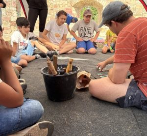 Eine Gruppe Kinder und Jugendlicher sitzt im Kreis und schaut zu wie eine erwachsene Person Mosaiksteinchen zusammensetzt. Die Person erklärt den Kids wie es geht.