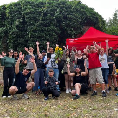 Die Teilnehmenden des Kunstcamps in Weißenfels vor einem Arbeit und Leben Pavillon. Sie reißen die Hände nach oben und jubeln,