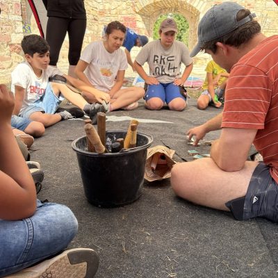 Eine Gruppe Kinder und Jugendlicher sitzt im Kreis und schaut zu wie eine erwachsene Person Mosaiksteinchen zusammensetzt. Die Person erklärt den Kids wie es geht.