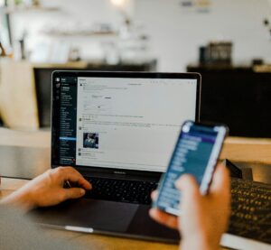 Man schaut einer Person über der Schulter, die in einem Cafe zu sitzen scheint. In der rechten Hand hat die Person ein Smartphone und zeitgleich tippt sie links etwas auf einem Laptop.