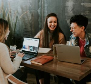 Drei Frauen sitzen an einem Tisch mit Laptops und Schreibutensilien. Sie unterhalten sich und lachen.