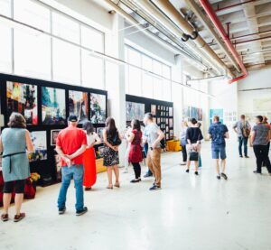 In einer Halle sind Bilder bzw. Kunstwerke ausgestellt. Personen schlendern durch die Halle oder stehen vor einige Bildern und betrachten sie.