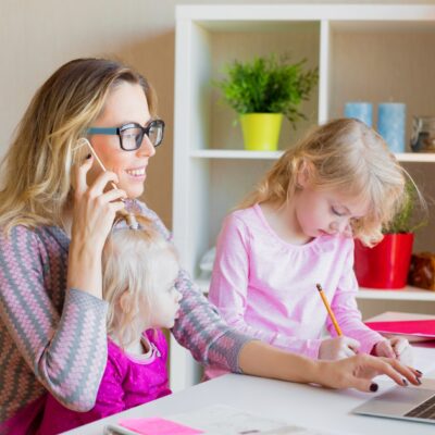 Eine Frau sitzt am Laptop und telefoniert. Ein Kind sitzt auf ihrem Schoß, ein zweites steht neben ihr und malt etwas.