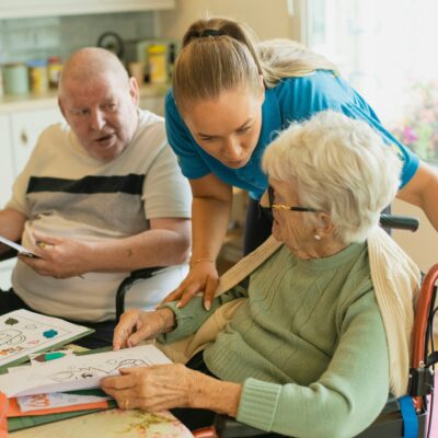 Ein älterer Mann in Rollstuhl sitzt mit einer Seniorin im Rollstuhl an einem Tisch. Eine Frau in Kleidung einer Pflegekraft steht zwischen ihnen und beugt sich zur Dame vor.