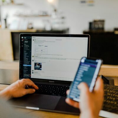 Man schaut einer Person über der Schulter, die in einem Cafe zu sitzen scheint. In der rechten Hand hat die Person ein Smartphone und zeitgleich tippt sie links etwas auf einem Laptop.