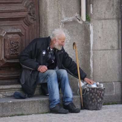Ein alter Mann sitzt auf einer Eingangstreppe, neben ihm ein Gehstock und ein Korb mit Pfandflaschen.