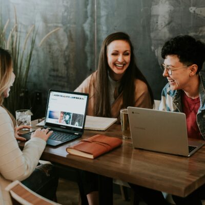 Drei Frauen sitzen an einem Tisch mit Laptops und Schreibutensilien. Sie unterhalten sich und lachen.