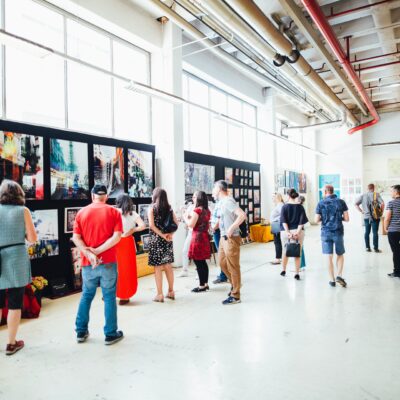 In einer Halle sind Bilder bzw. Kunstwerke ausgestellt. Personen schlendern durch die Halle oder stehen vor einige Bildern und betrachten sie.