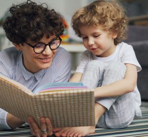 Eine Frau liegt auf dem Boden und hat ein geöffnetes Buch in der Hand. Neben ihr sitz ein Kleines Kind und zeigt Sachen in dem Buch. Beide lächeln.