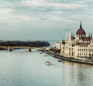 Blick auf einen breiten Fluss, im Hintergrund eine Brücke, auf der rechten Seite ein altes Gebäude, das das Parlament von Ungarn ist.