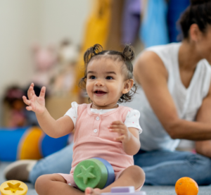 Blick in eine Kita. Mehrere Kinder und einer Erzieherin sitzen auf dem Boden und Spielen. Ein kleines Mädchen im Vordergrund klatscht.