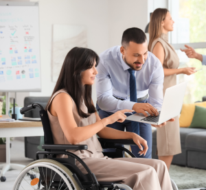 Ein Mann beugt sich mit einem Laptop zu einer Frau herunter. Diese sitzt im Rollstuhl und zeigt auf den Bildschirm.