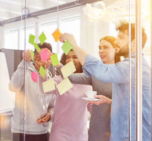 Man Blcikt durch eine gläserne Wand, an der auf der anderen Seite Post-Its kleben. Vier Personen stehen und sortieren sie um.
