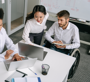An einem Konferenztisch sitzt eine Frau mit Laptop, neben ihr Seht eine weitere Frau, die am Laptop etwas zeigt. Sie zeigt es einem Mann im Rollstuhl der in einem Buch Notizen macht.