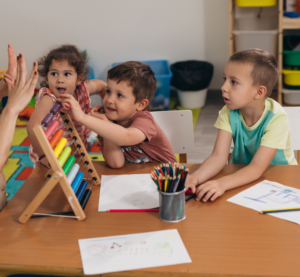 Eine Frau steht an einem Tisch und zeigt mit den Fingern Zahlen. Auf dem Tisch steht ein bunter Rechenschieber. Drei Kinder versuchen die Kugeln hin und herzuschieben.