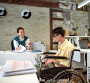 Ein Mann im Rollstuhl hat einen Laptop auf dem Schoß. Er spricht mit einer Frau, die hinter ihm an einem Schreibtisch sitzt und Unterlagen in der Hand hat.