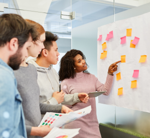 An einer Glaswand hängt ein Plakat mit mehreren Klebezetteln. eine Frau zeit auf einen Klebezettel und erklärt den drei Personen hinter ihr etwas.