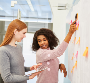 Eine Frau steht an einem Plakat und zeigt mit dem Stift auf einen Klebezettel. Sie schaut sich fragend um auf das Tablet der Frau hinter ihr.