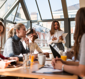 Mehrere Personen sitzt an einem Konferenztisch. Es liegen verschiedene Unterlagen auf dem Tisch sowie einige Getränke stehen dort. Vorn steht eine Frau mit Unterlagen in der Hand und erklärt etwas.