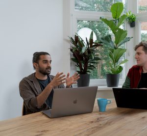 Zwei Männer mit jeweils einem Laptop sitzen an einem Tisch und unterhalten sich.
