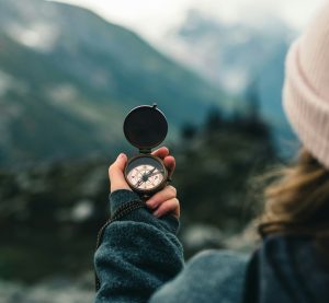Man blickt einer Frau über die Schulter, sie hält einen Kompass in der Hand und blickt über diesen in die Berge.