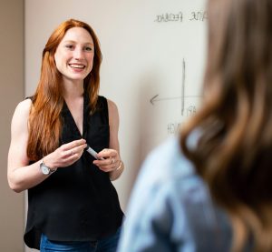Wir schauen über die Schulter einer Frau und blicken auf eine anderer Frau, die mit einem Marker vor einem Whiteboard steht.