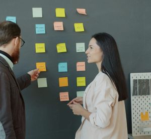 Zwei Personen stehen vor einer Wand mit mehreren Klebezetteln. Der Mann zeigt mit einem Stift auf einen der Zettel.