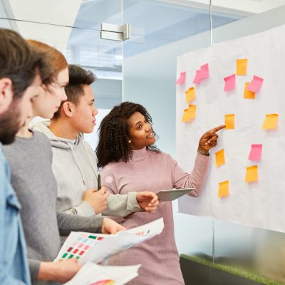 An einer Glaswand hängt ein Plakat mit mehreren Klebezetteln. eine Frau zeit auf einen Klebezettel und erklärt den drei Personen hinter ihr etwas.
