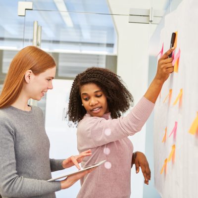 Eine Frau steht an einem Plakat und zeigt mit dem Stift auf einen Klebezettel. Sie schaut sich fragend um auf das Tablet der Frau hinter ihr.