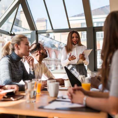 Mehrere Personen sitzt an einem Konferenztisch. Es liegen verschiedene Unterlagen auf dem Tisch sowie einige Getränke stehen dort. Vorn steht eine Frau mit Unterlagen in der Hand und erklärt etwas.