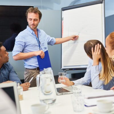 Ein Mann steht vor einer Tafel und zeit etwas darauf. Mehrere Personen sitzen um einen Tisch und betrachten die Tafel.