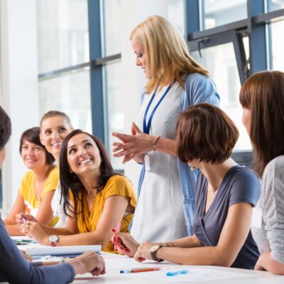 Mehrere Frauen sitzen an einem Tisch und blicken zu einer, die sich hingestellt hat und etwas erklärt.