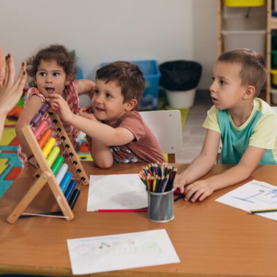 Eine Frau steht an einem Tisch und zeigt mit den Fingern Zahlen. Auf dem Tisch steht ein bunter Rechenschieber. Drei Kinder versuchen die Kugeln hin und herzuschieben.