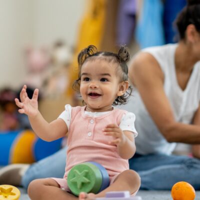 Blick in eine Kita. Mehrere Kinder und einer Erzieherin sitzen auf dem Boden und Spielen. Ein kleines Mädchen im Vordergrund klatscht.