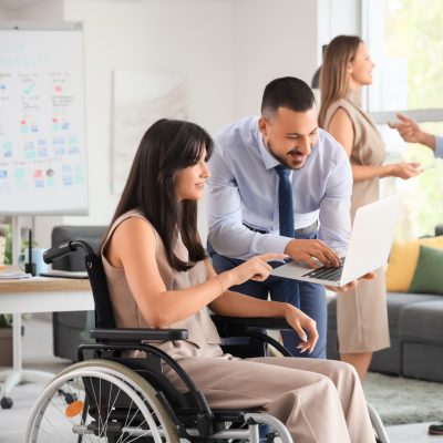 Ein Mann beugt sich mit einem Laptop zu einer Frau herunter. Diese sitzt im Rollstuhl und zeigt auf den Bildschirm.