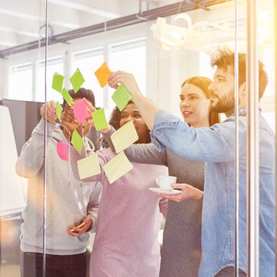 Man Blcikt durch eine gläserne Wand, an der auf der anderen Seite Post-Its kleben. Vier Personen stehen und sortieren sie um.