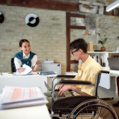Ein Mann im Rollstuhl hat einen Laptop auf dem Schoß. Er spricht mit einer Frau, die hinter ihm an einem Schreibtisch sitzt und Unterlagen in der Hand hat.